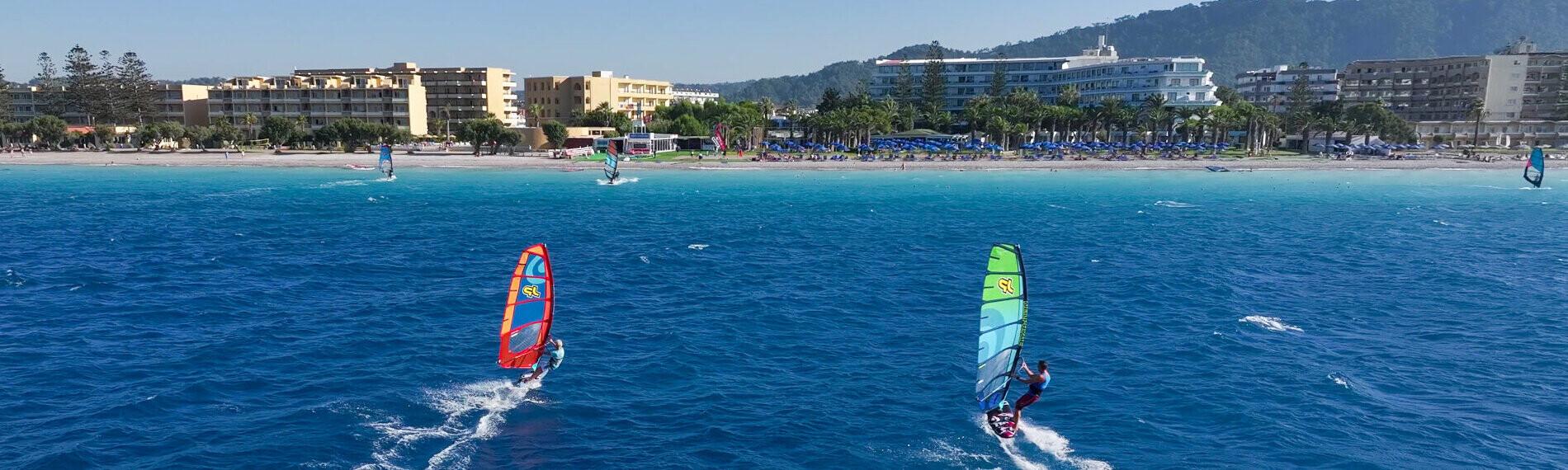 Windsurfer gleiten über türkises Meer vor Küstenlinie Rhodos mit Hotels.