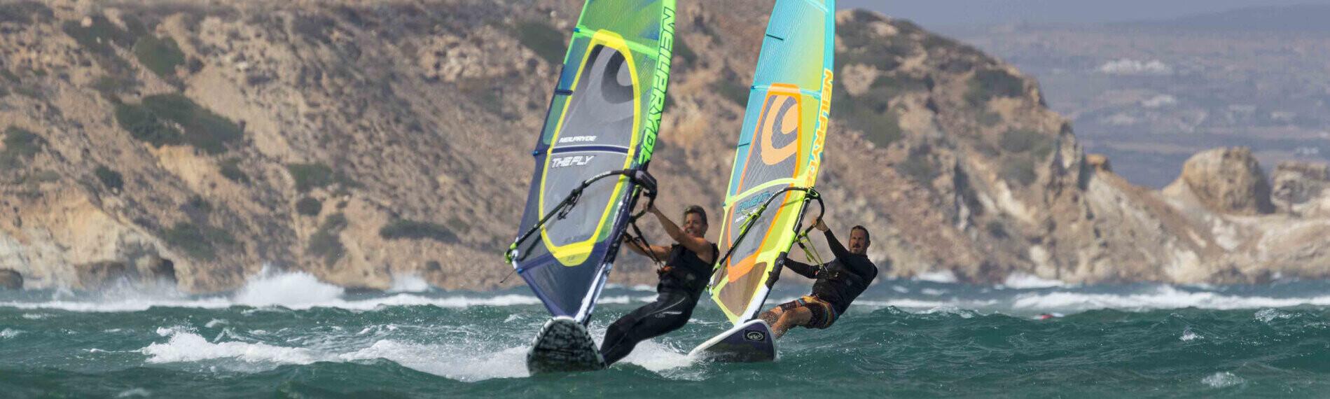 Zwei Windsurfer auf welligem Meer vor felsiger Küste Naxos