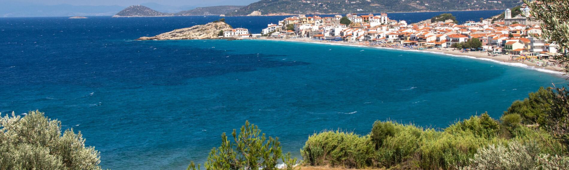 Samos - Kokkari - Küstenstadt mit blauem Meer und Bergen im Hintergrund, umgeben von grüner Vegetation unter klarem Himmel.