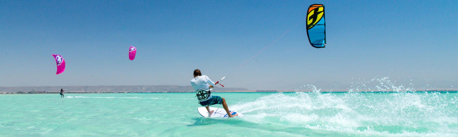 Kitesurfer in Ras Soma, Ägypten auf türkisem Wasser mit bunten Schirmen im Himmel.