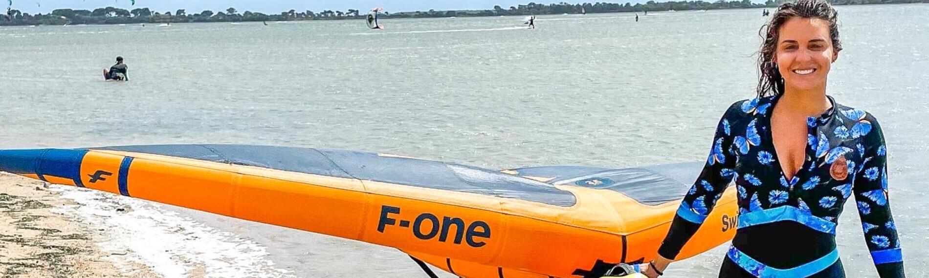 Sizilien - KBC, Wingfoil Frau läuft mit Wingfoil-Ausrüstung am Strand.